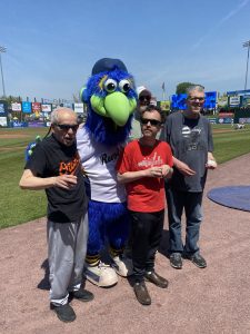 3 men and the York Revolution baseball team mascot standing on a baseball field together.